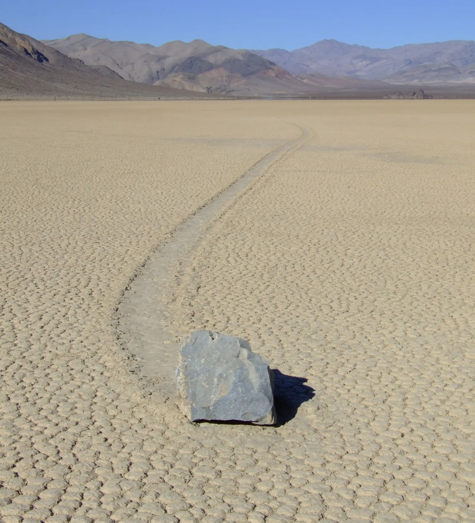 A stone on a cracked dry lakebed with a long track trailing behind it, Death Valley National Park, California
