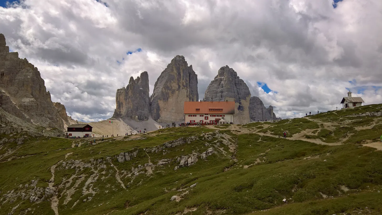 A stone mountain hut on a grassy saddle facing three rock towers, Dolomites, Italy
