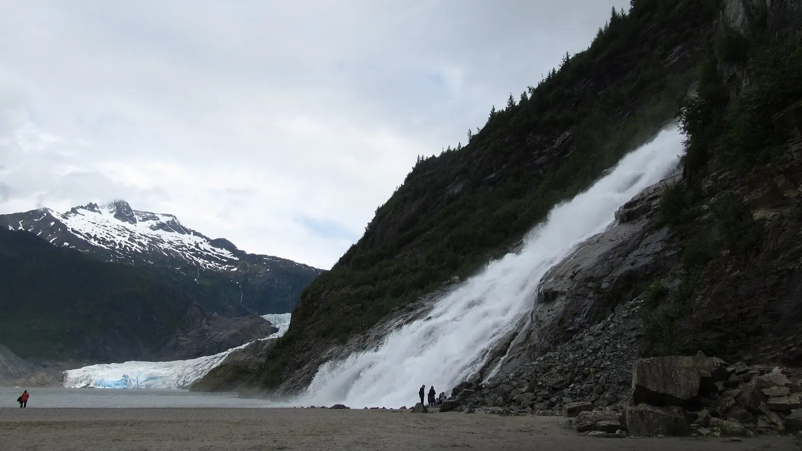A tall waterfall pouring onto a gravel bar beside a lake with a glacier nearby, Juneau, Alaska