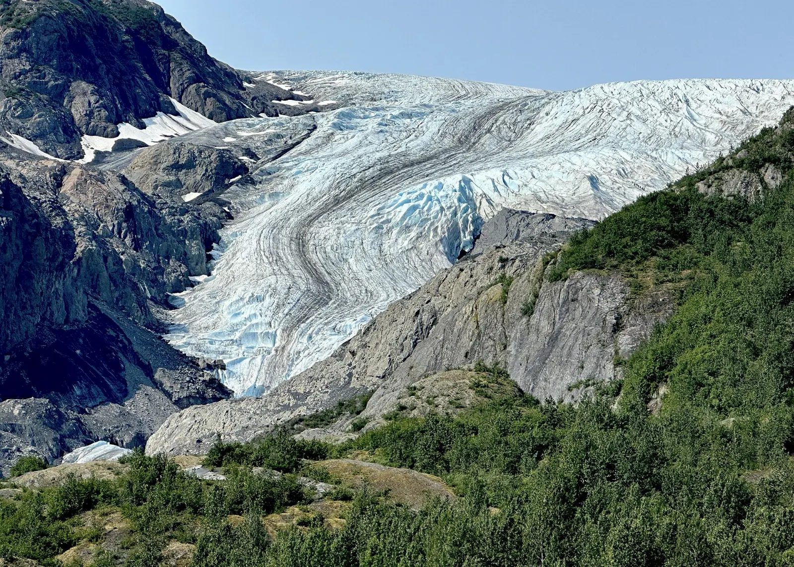 A blue and white glacier tongue descending a rocky valley between green slopes, Kenai Fjords National Park, Alaska