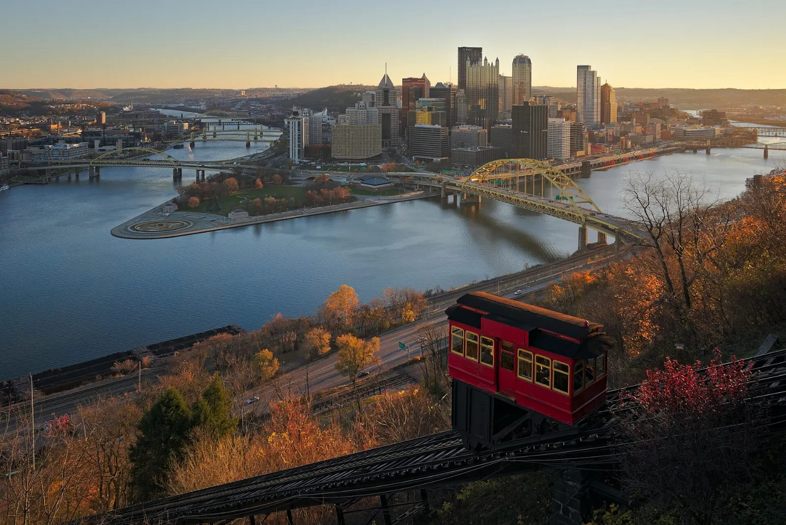 A small red funicular car climbing a steep track up a wooded hillside above a river, Pittsburgh, Pennsylvania